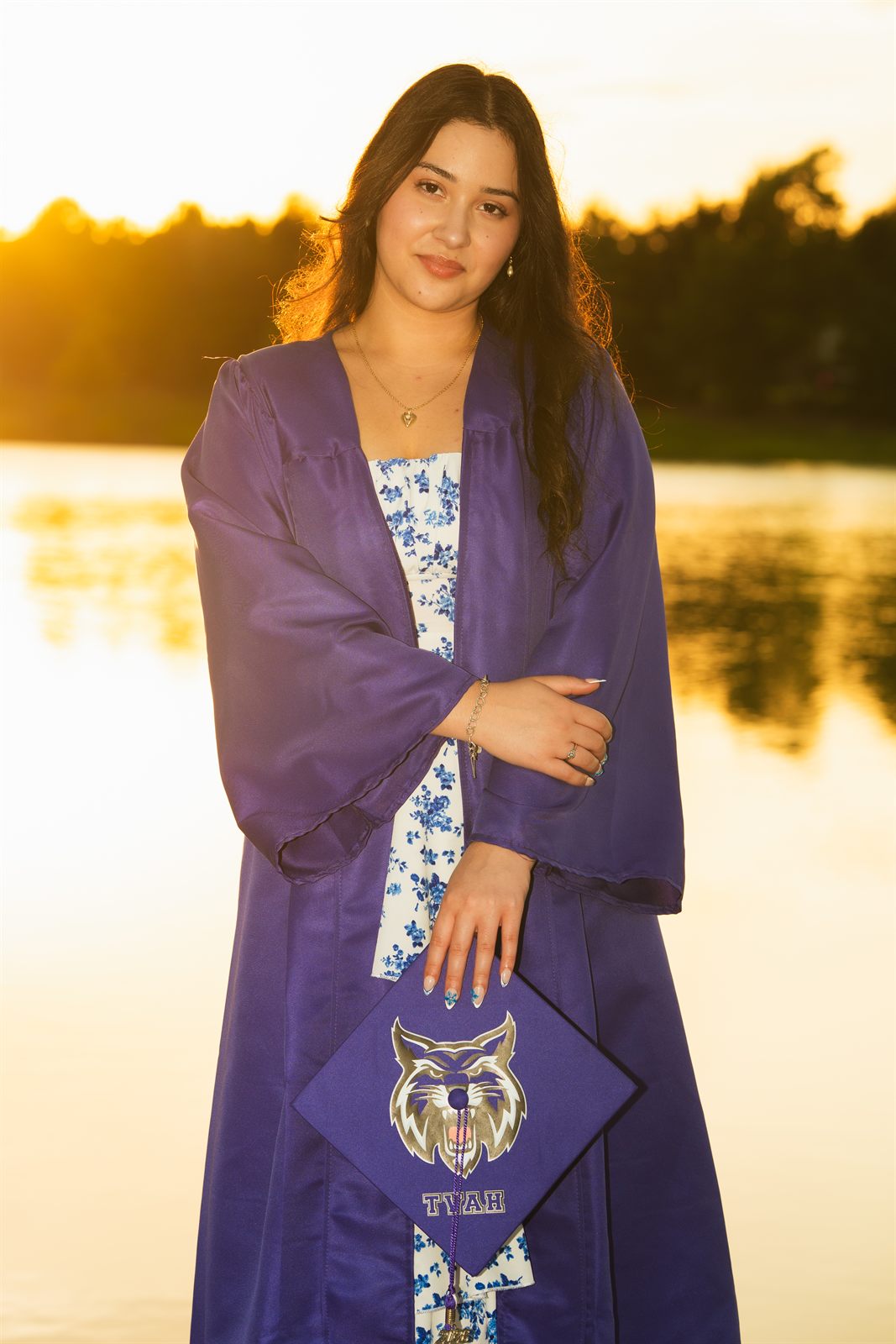 Graduate in a purple gown holding her cap at a sunset lake