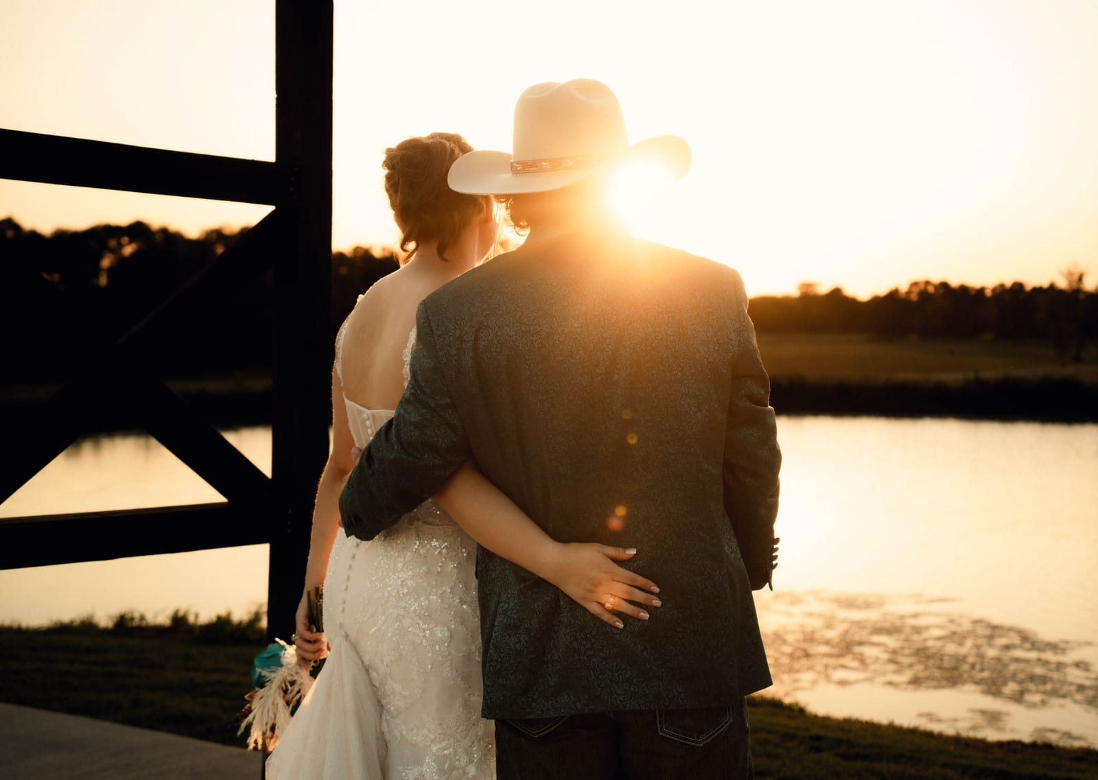 Bride and groom in a cowboy hat holding each other beside a lake at sunset