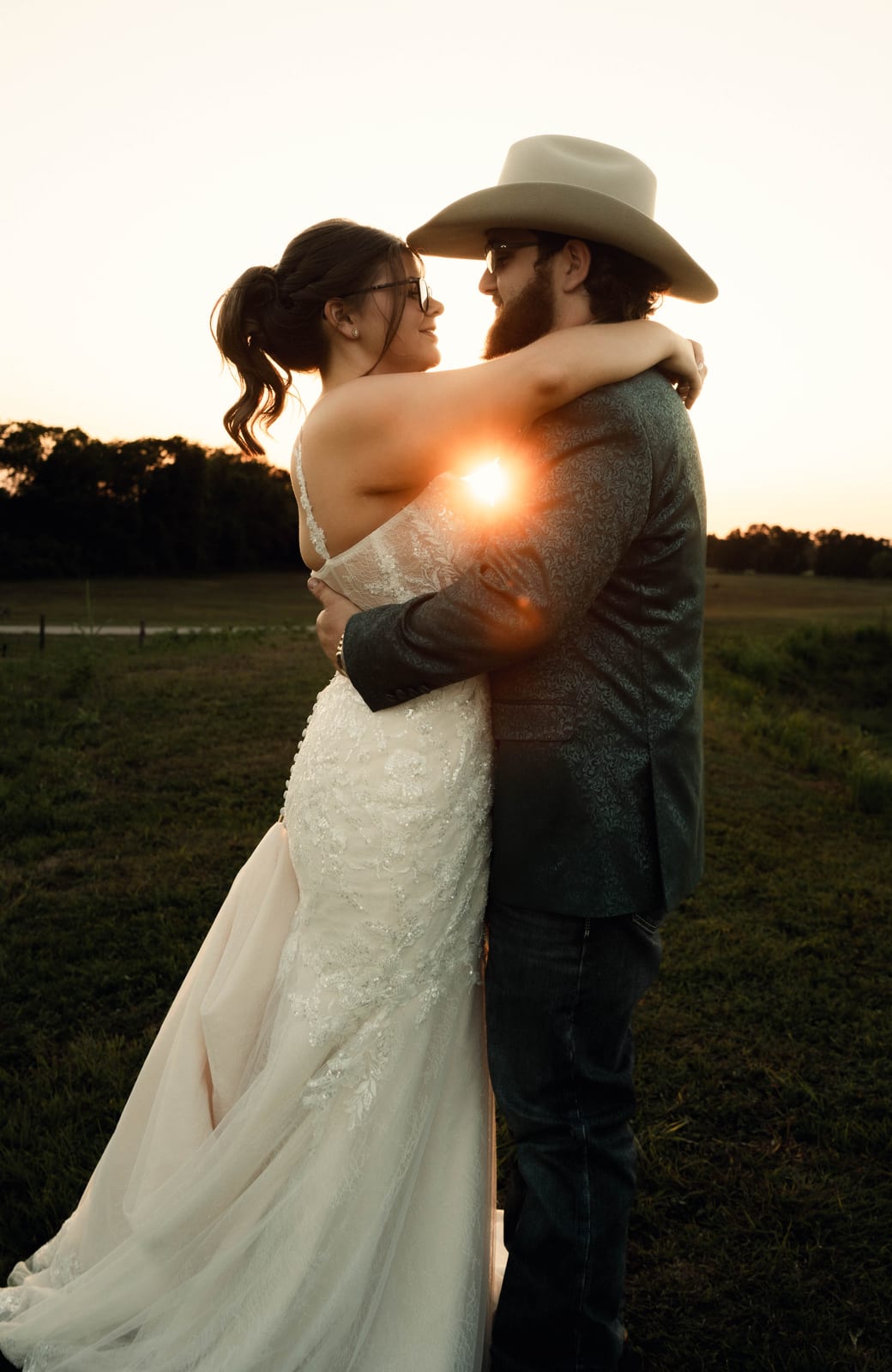 Bride and groom in a cowboy hat embracing in an open field as the setting sun flares between them