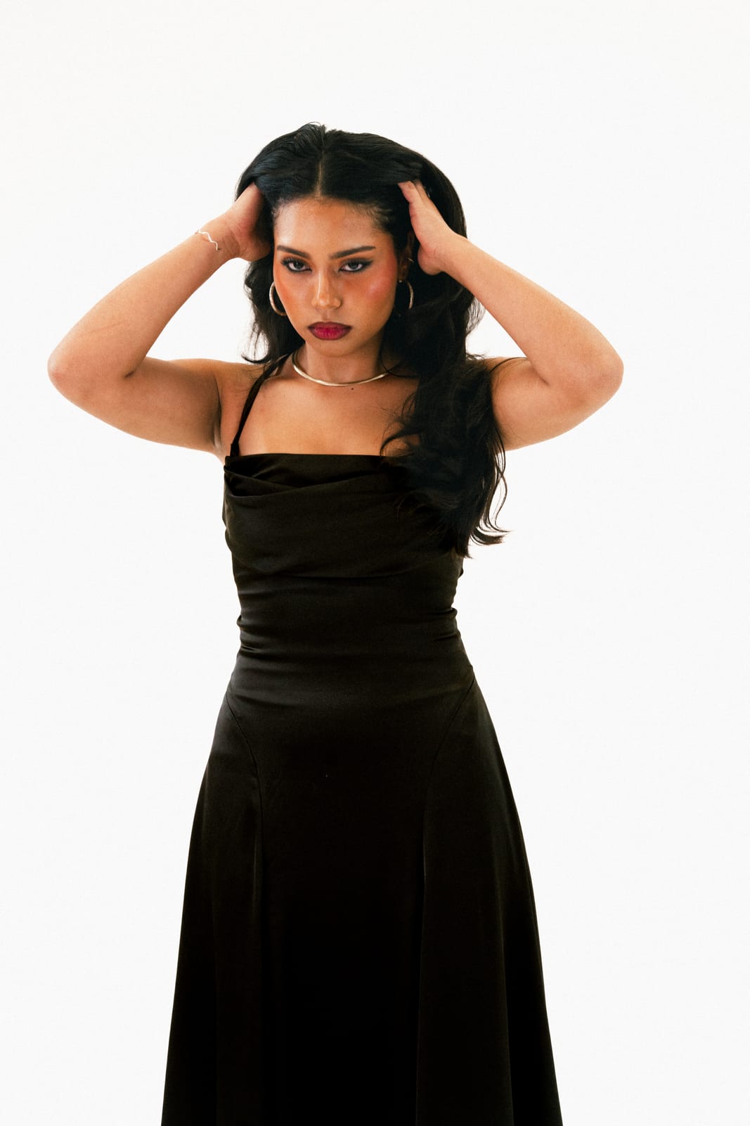 Woman in a black slip dress with her hands in her hair against a white backdrop