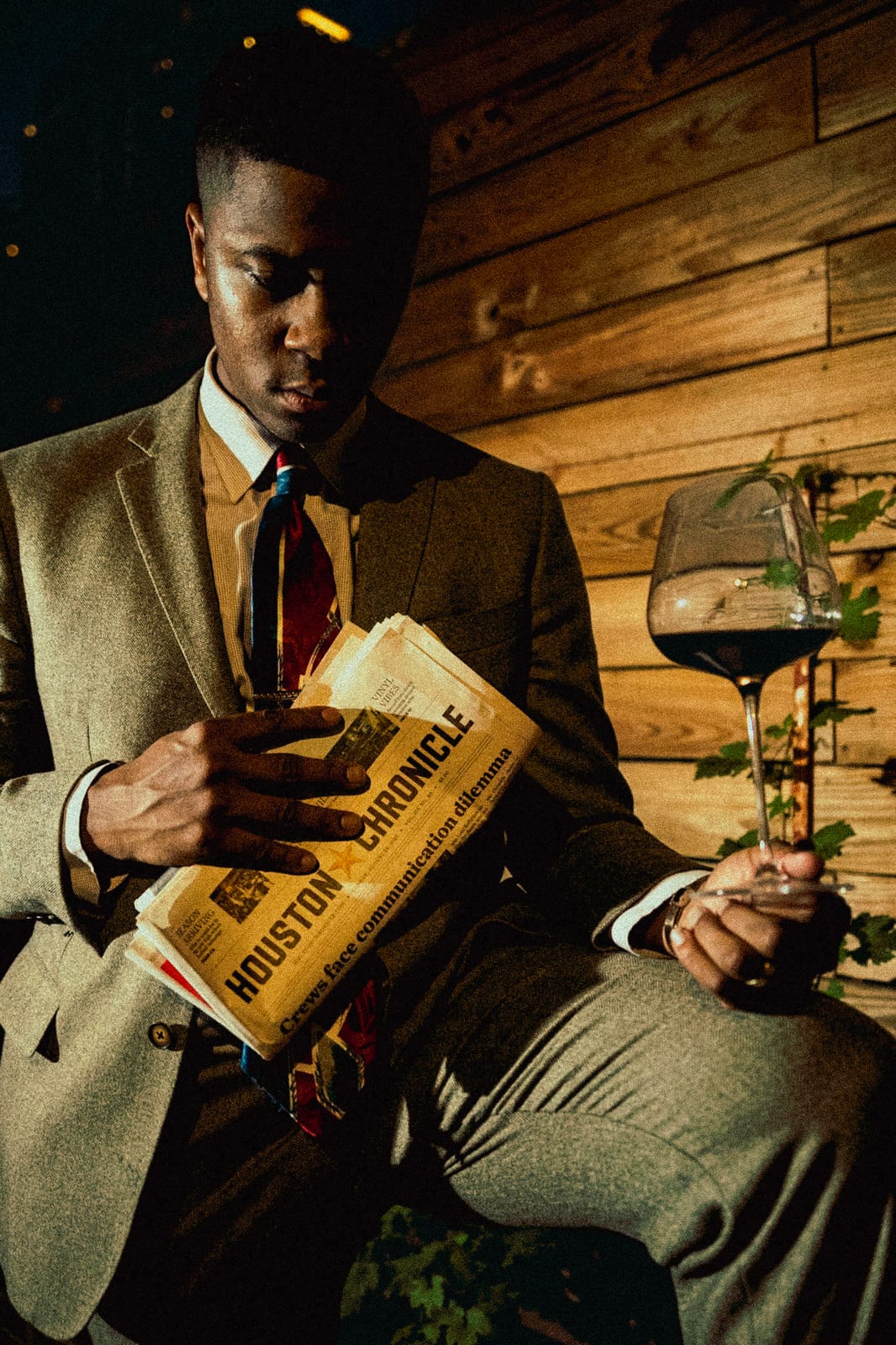 Man in a tweed suit reading the Houston Chronicle with a glass of red wine against a wood wall