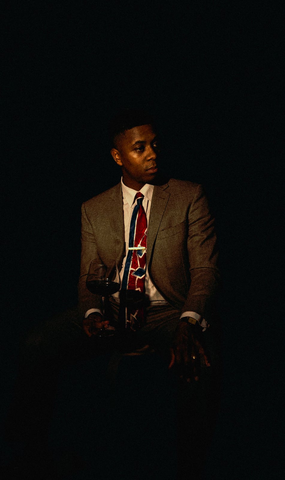 Man in a tweed suit and red tie seated in deep shadow with a glass of wine