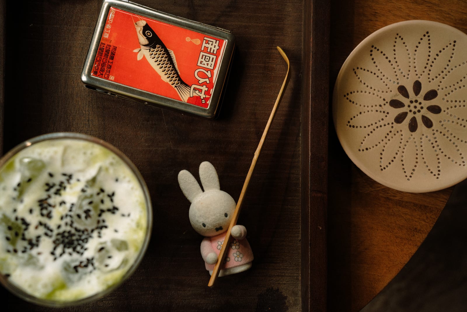 Overhead view of a green drink topped with black sesame beside a vintage koi tin and a small rabbit figurine