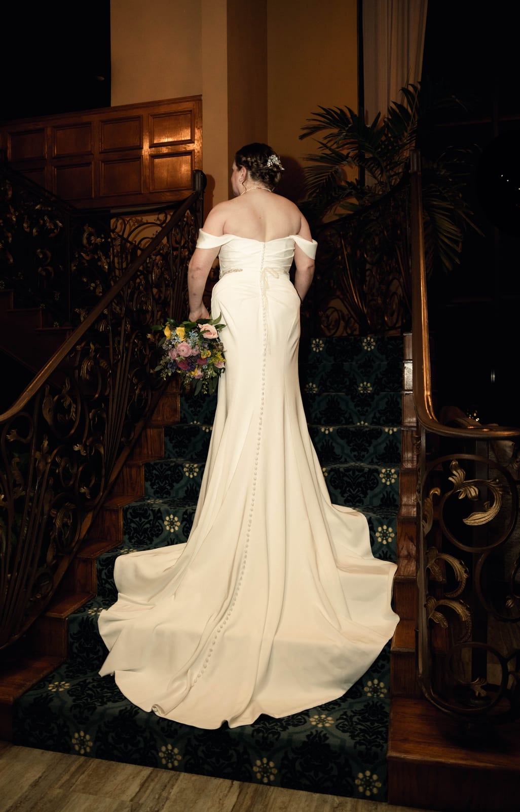 Bride in an off-the-shoulder gown, her long train spread across a carved wooden staircase