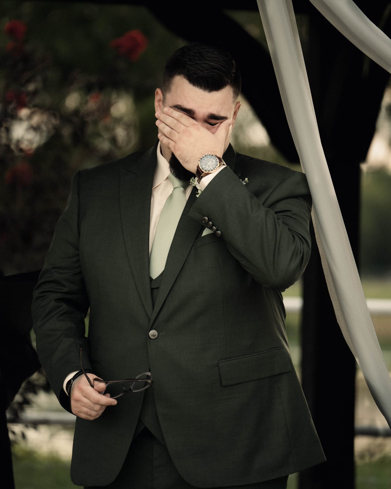 Groom in a dark green suit with his hand over his eyes, holding his glasses